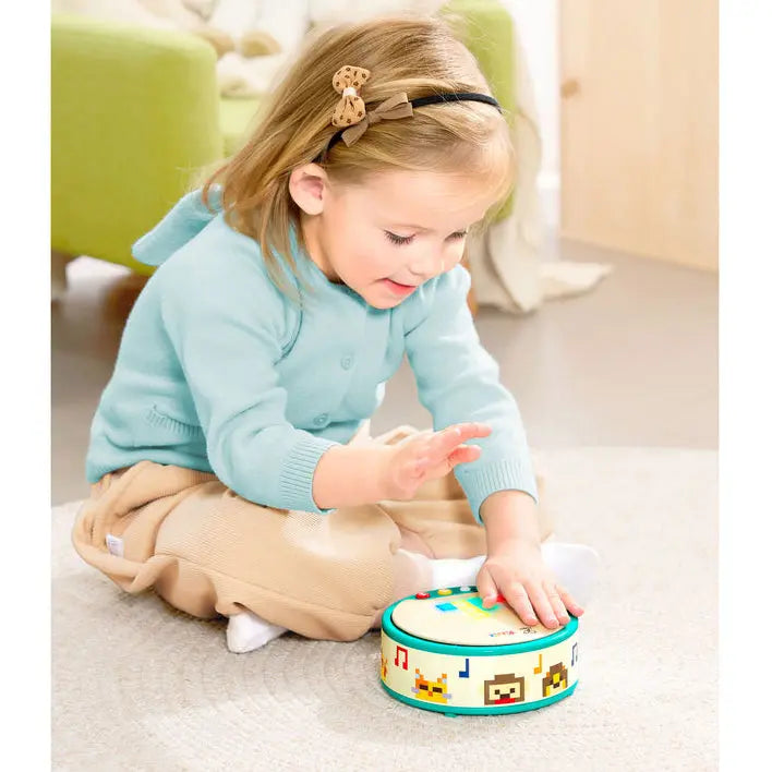 A young child sitting on a light-colored carpet in a bright room, playing with a small round toy drum. The child is wearing a light blue cardigan and tan pants, with a stuffed animal and a green chair visible in the background.