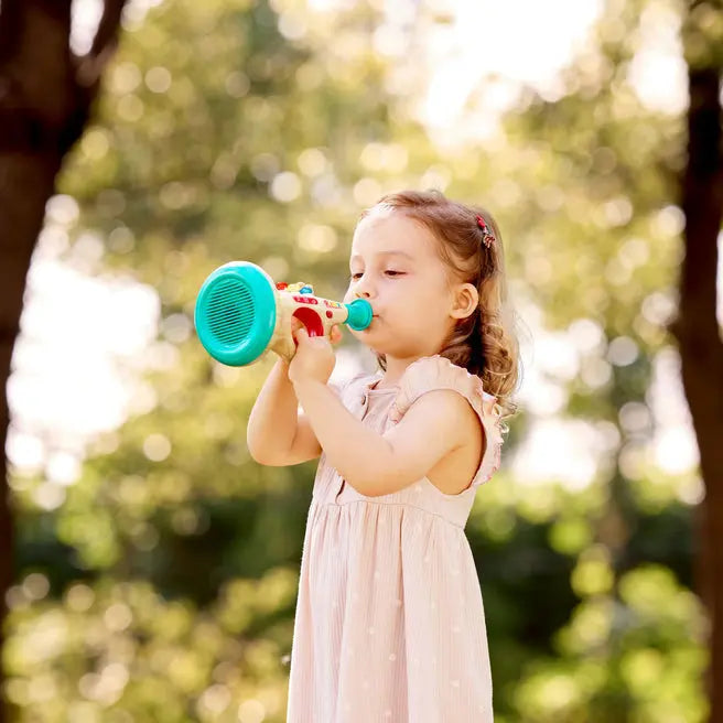 A young girl in a light pink dress playing with a small teal and red toy horn outdoors in a sunlit park with blurred trees.