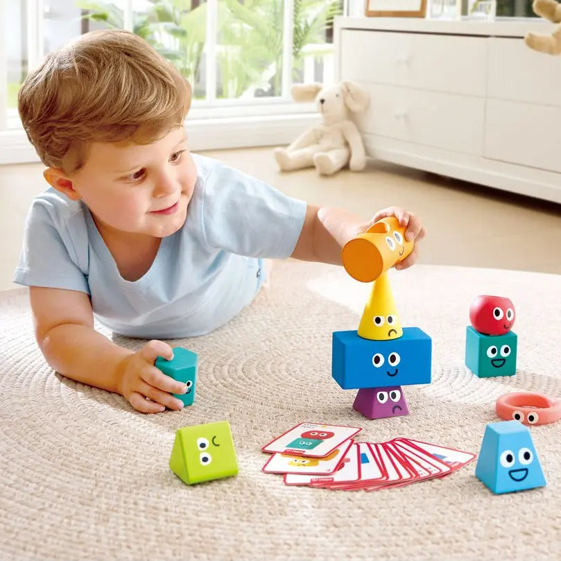 A young child lies on a light beige rug in a bright room, playing with colorful wooden toy blocks and cards. The toys include blue, green, yellow, red, and pink geometric shapes with cartoon faces, along with small cards.