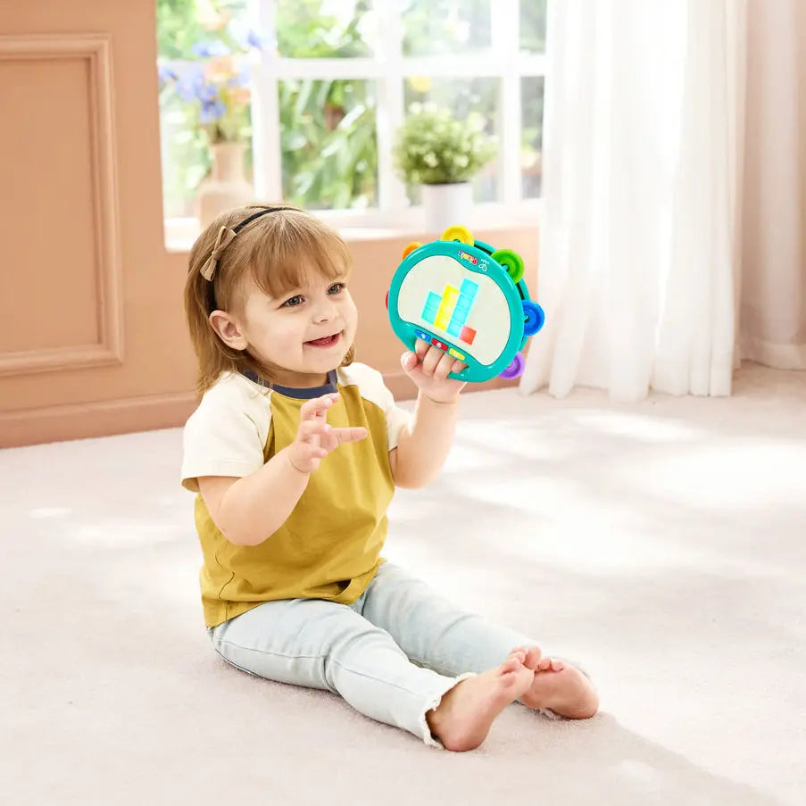 A young child sitting on a light carpet in a bright room, holding a colorful, sound-reactive toy rattle. The toy has a turquoise-blue outer ring with colorful tabs and a white center.