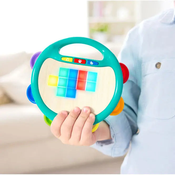A child in a light blue button-up shirt holds a colorful wooden toy rattle shaped like a steering wheel in a bright, softly blurred living room setting.