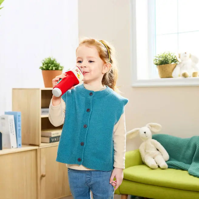 A young girl stands in a bright children's room wearing a teal knit vest over a cream shirt and blue jeans, holding a red cup. A green sofa with a yellow star pillow and a white stuffed rabbit is visible in the background.